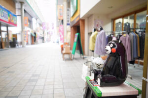 八代本町アーケードの商店街の風景。婦人服店の店先でお客さんを待っているような、もの思いにふけるくまモン。