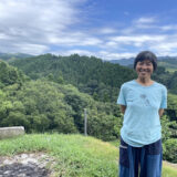 熊本県御船町の豊かな森を背景に、にこやかに立つ國武林業の創業者・國武信子さん。