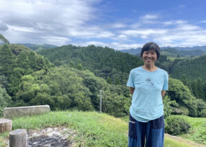 熊本県御船町の豊かな森を背景に、にこやかに立つ國武林業の創業者・國武信子さん。