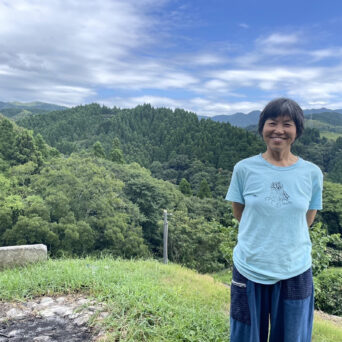 熊本県御船町の豊かな森を背景に、にこやかに立つ國武林業の創業者・國武信子さん。