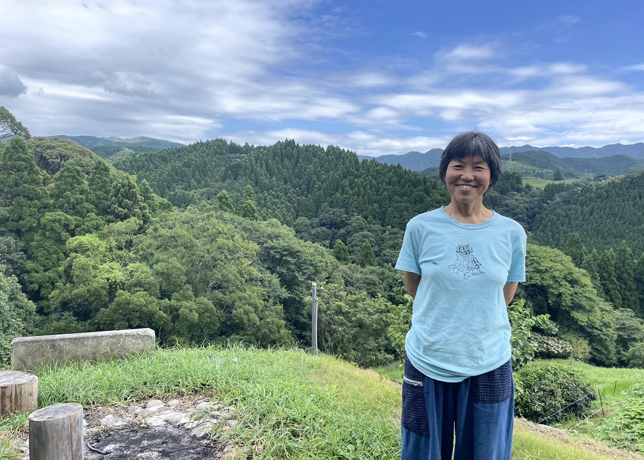 熊本県御船町の豊かな森を背景に、にこやかに立つ國武林業の創業者・國武信子さん。