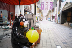 八代本町アーケードの散策中に見つけた写真スポット。赤い和傘の下で、八代名物の晩白柚（ばんぺいゆ）を大事そうに抱えるくまモン。