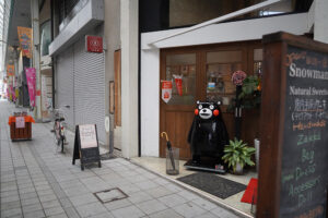 八代本町アーケードのおしゃれな雑貨店の店先。看板の横に立って、笑顔で来店者を出迎えてくれるようなくまモン。