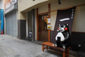 八代本町アーケードの路地裏にある飲食店（居酒屋）の店先。大きなおにぎりを抱えてベンチに座る、食いしん坊なくまモンが可愛い。