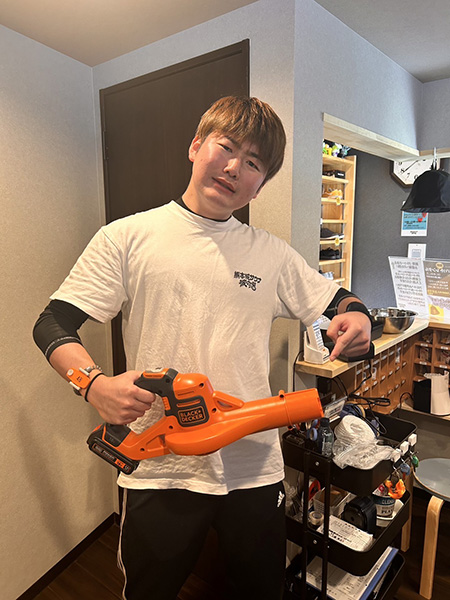 熊本の熱波師しげちゃんが、熱波サービスで使用するオレンジ色のブロワーを手に持ち、笑顔で紹介している。