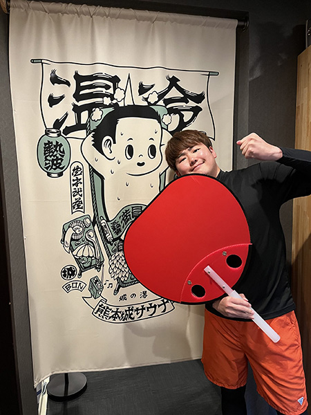 熊本城サウナののれんの前で、熱波師しげちゃんがパフォーマンスで使う大きな赤い熱波うちわを持ってポーズをとっている。