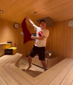 熊本城サウナ城の湯の木製サウナ室で、アウフギーサーのしげちゃんが赤いタオルをダイナミックに振ってアウフグースのパフォーマンスをしている様子。