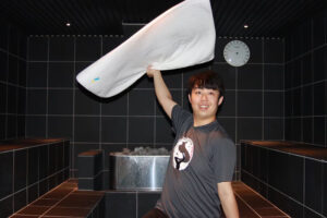 黒いタイル張りのモダンなサウナ室で、アウフギーサーのKCが白いタオルを大きく広げてアウフグースのパフォーマンスをしている様子。