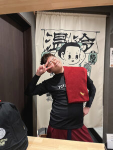 熊本城サウナののれんの前で、アウフギーサーのしげちゃんが黒いTシャツを着て、首にかけた赤いタオルを持ちながらピースサインをしている。