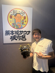 熊本城サウナ城の湯のロゴの前で、アウフギーサーのしげちゃんが笑顔でシーサーのような置物を指さしている。