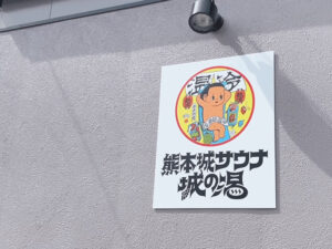 熊本城サウナ城の湯の建物の外壁に取り付けられた看板のクローズアップ。「温冷浴」のキャラクターイラストと施設のロゴがデザインされている。