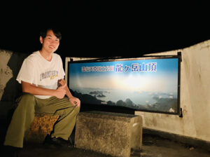 雲仙天草国立公園の龍ヶ岳山頂の看板の前で笑顔でサムズアップする高校生。天草の絶景ポイントを示す夜間の写真。