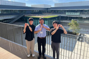 商品開発チームのメンバーと思われる3人の男性が、近代的な建物を背景に笑顔でポーズをとっている。チームワークの良さや、研究開発の明るい雰囲気が伝わる一枚。